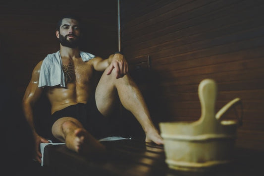 A man sitting on a bench in a sauna with a wooden bucket next to him.