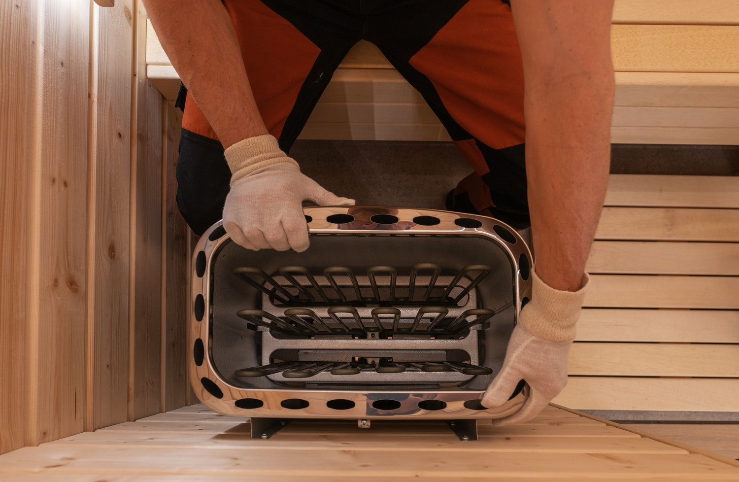 A person wearing orange and black work pants and white gloves is installing a stainless steel heater in a wooden room. The person is arranging the heater within a rectangular opening between wooden panels. The room appears to be part of a sauna.