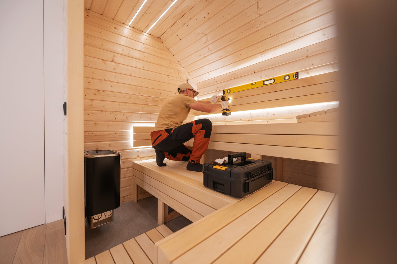 A person in a beige shirt and orange pants is working inside a wooden sauna. They are using a level tool, drilling into the backrest. The sauna has wooden benches and walls, with a toolbox set on one of the benches. A heater is positioned in a corner.